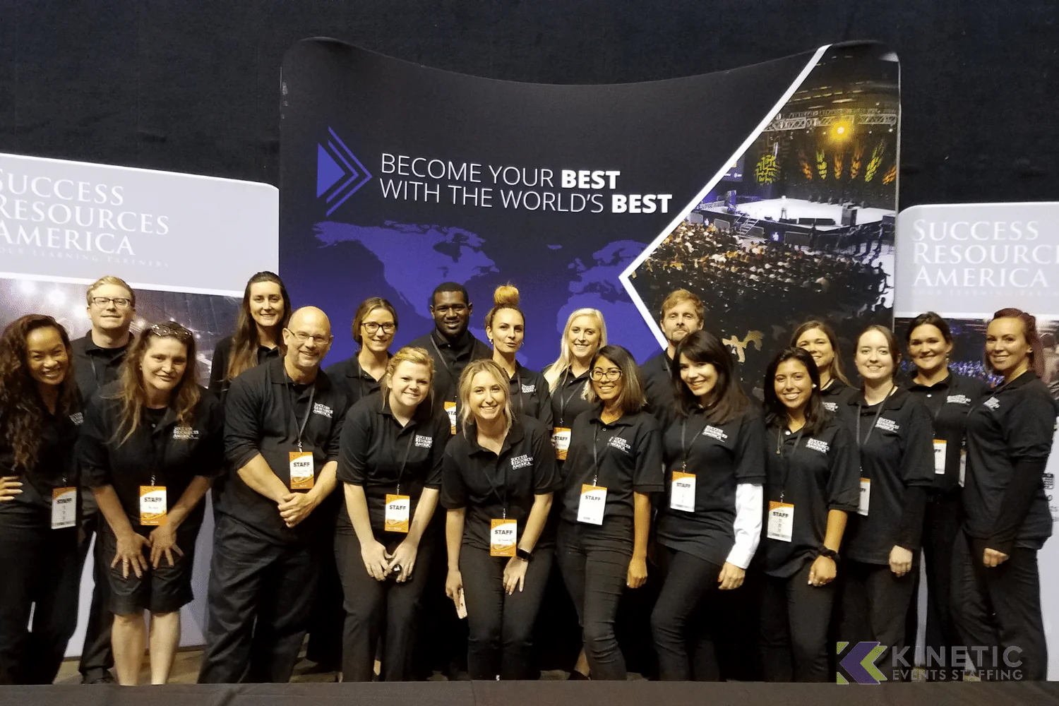 Group of 14 people in black shirts with 'STAFF' lanyards stand in front of a backdrop. | SUCCESS RESOURCES AMERICA OUR LEARNING PARTNERS STAFF STAFF BECOME YOUR BEST WITH THE WORLD'S BEST SUCCESS RESOURC AMERICA STAFF STAFF STARR STAFF KINETIC KK EVENTS STAFFING